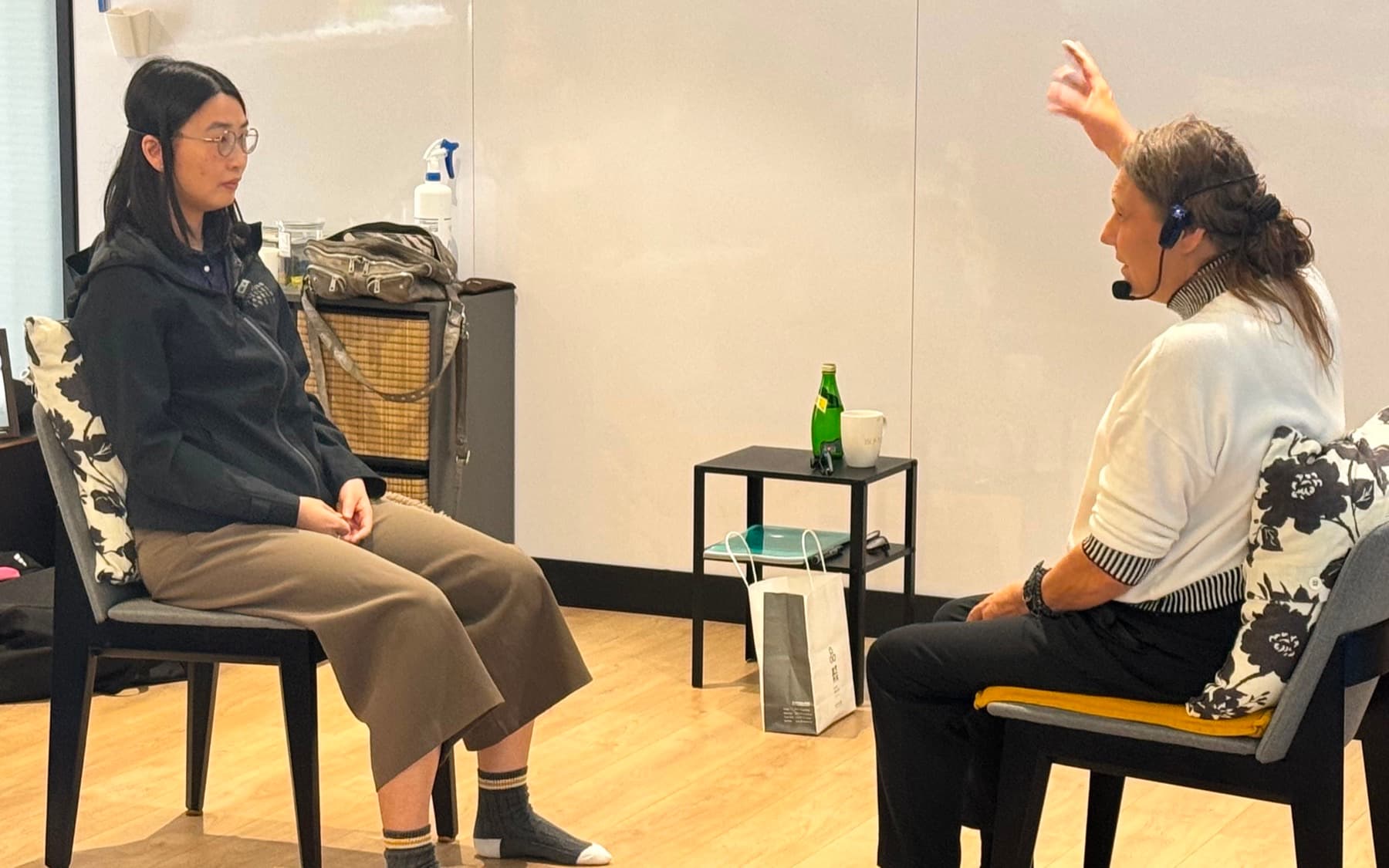 Two people sitting on chairs facing each other in a bright room, one speaking and gesturing while the other listens, attention on how the exchange is landing in the body.