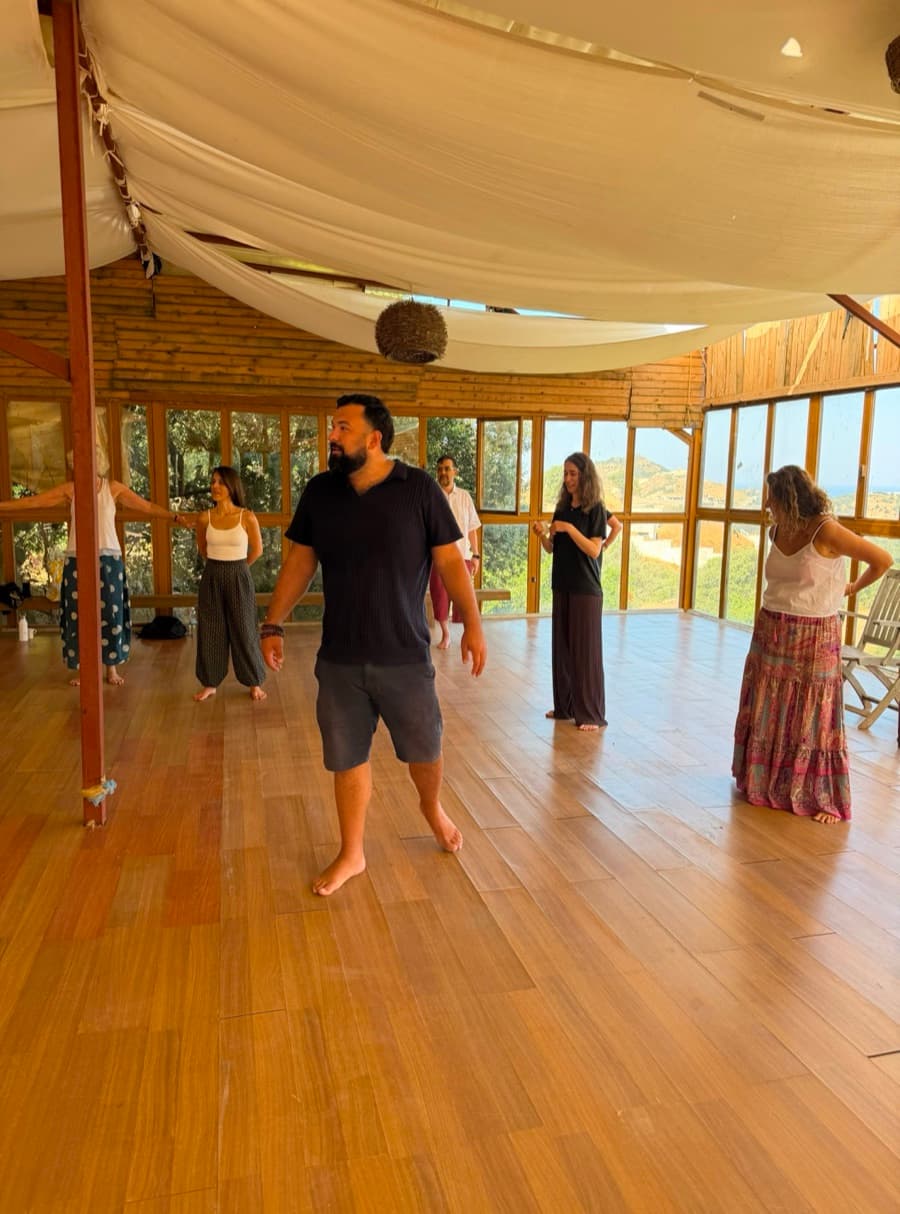 A group of participants standing barefoot in a hillside studio, working through a movement exercise.