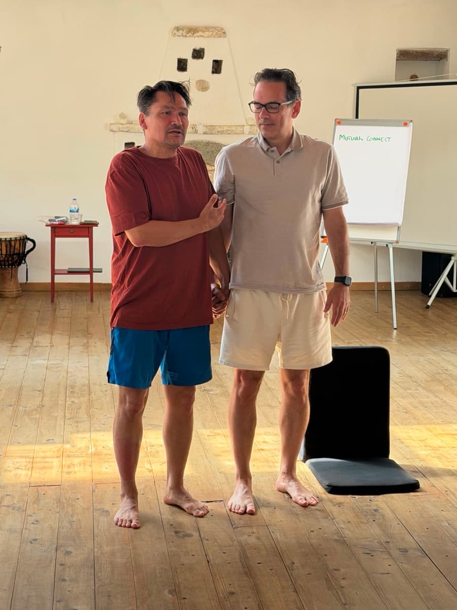 Two participants standing side by side in a training room, one supporting the other's arm, with “Mutual Connect” written on the whiteboard behind them.